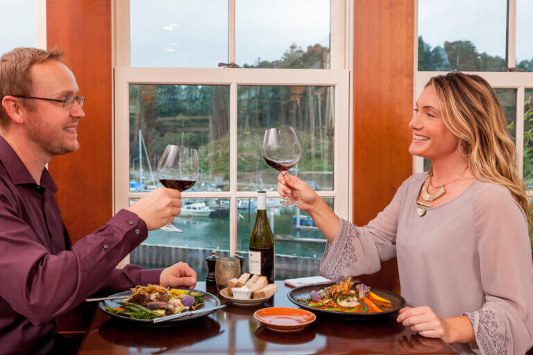 Young couple eating dinner indoors overlooking the Noyo River and toasting wine glasses